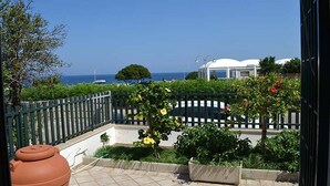 Un balcón con vista al mar, un jardín con plantas en flor y un edificio al fondo.