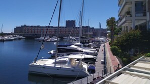 Un port de plaisance où sont amarrés de nombreux yachts, un bâtiment avec des balcons en arrière-plan et un ciel bleu dégagé.