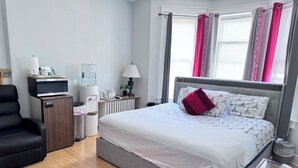 A modern hotel room with a grey bed, a black chair, a minibar, a water dispenser, and a radiator.