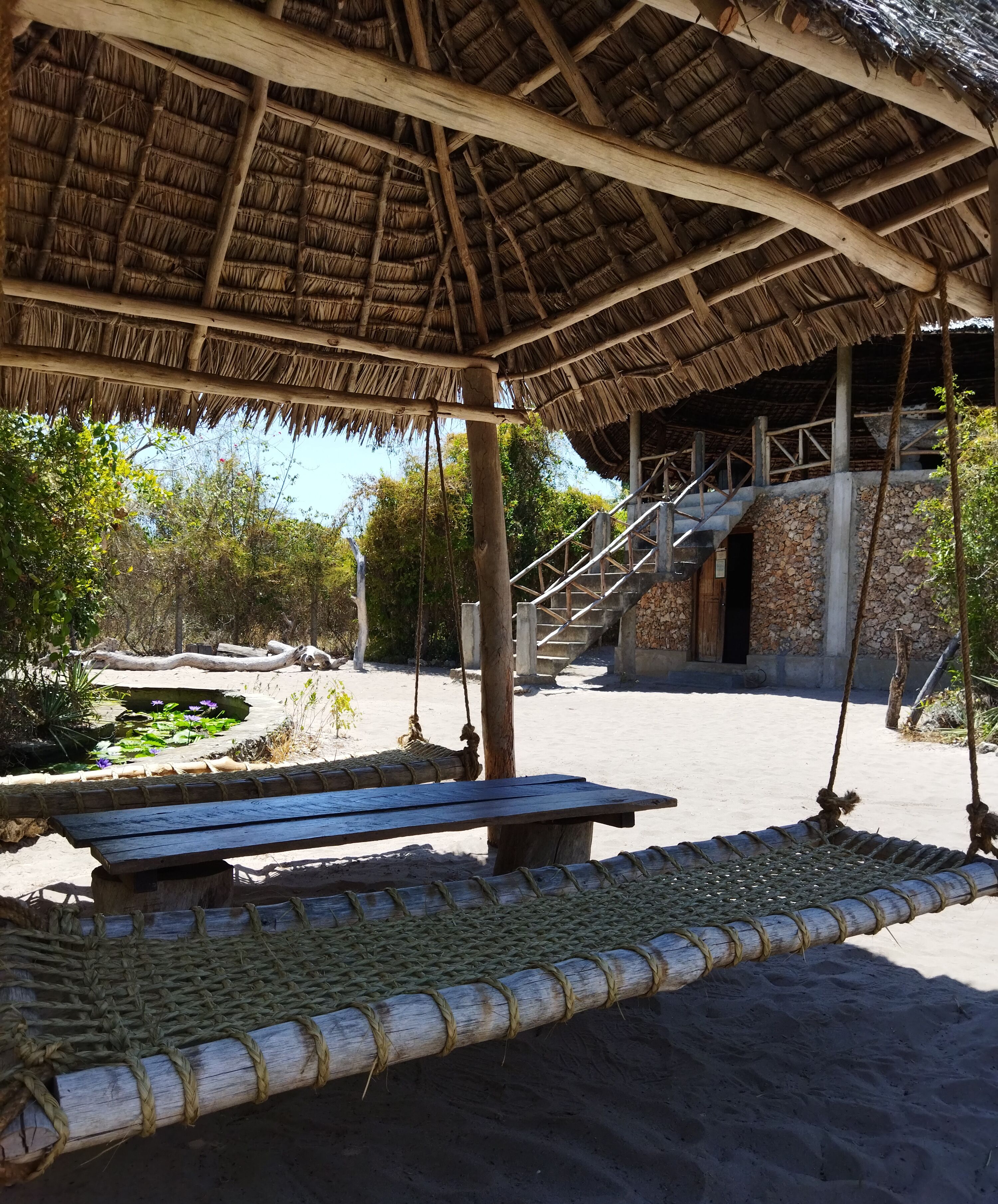 A thatched-roof structure with a wooden bench, a stone wall, and a staircase leading to an upper level.