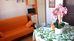 A room with an orange sofa, a small table with a floral tablecloth, a vase with pink flowers, and a painting on the wall. - Holiday Home With Balcony and air Conditioning in Torre Dell'o (Melendugno)