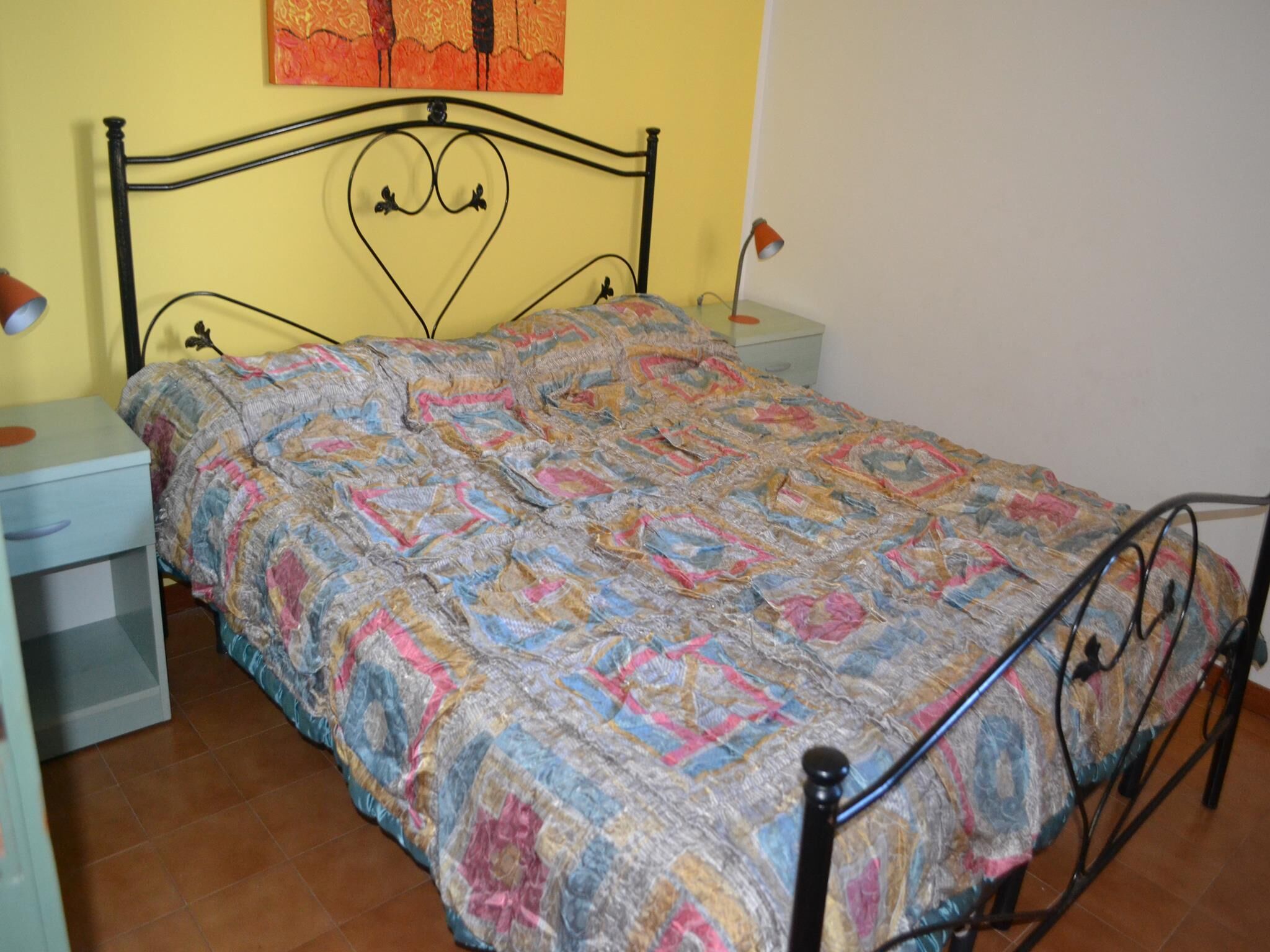 Ein Bett mit gemustertem Bettbezug, ein Nachttisch mit Lampe und eine Wand mit einem Gemälde.