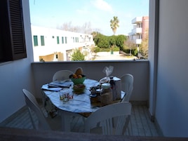 A balcony with a table set for dining, a bowl of fruit, and a view of buildings and trees.