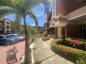 A street view with parked cars, a bench, palm trees, and a building with a gated entrance. - Spacious 3-bedroom Apartment in Santo Domingo! (Santo Domingo Norte)