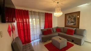 A modern living room with a grey sectional sofa, red cushions, a red rug, and a chandelier. - Spacious 3-bedroom Apartment in Santo Domingo! (Santo Domingo Norte)