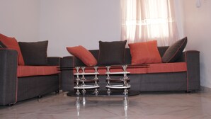 A modern living room with a grey sofa, red cushions, and a glass coffee table with metal legs. - Elu Hotel Studio - Bonamoussadi (Douala)