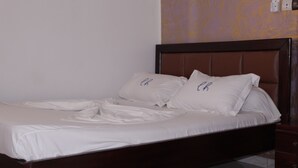 A neatly made bed with white linens and two pillows, a wooden headboard, and a wall with a patterned wallpaper. - Elu Hotel Studio - Bonamoussadi (Douala)