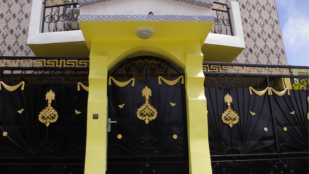 A building with a yellow facade and black gates featuring ornate golden designs. - Elu Hotel Studio - Bonamoussadi (Douala)