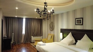 Une chambre d’hôtel avec un grand lit, un lustre, une chaise, un bureau et une fenêtre avec des rideaux.