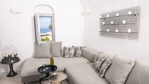 A modern living room with a grey sofa, a glass coffee table, a wine bottle, and a view of the sea through an open window. - Executive Villa With Private Outdoor Plunge-pool, Sea Sunset View 4 Guests (Santorini)