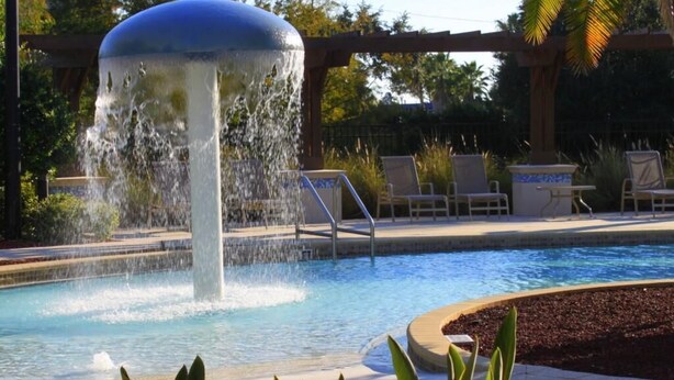 A swimming pool with a water fountain, surrounded by lounge chairs and palm trees.