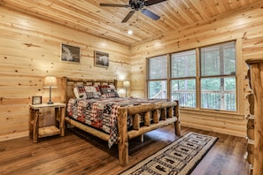 A log cabin bedroom with a wooden bed, a nightstand, a ceiling fan, and large windows.