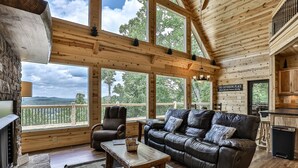 L’intérieur chaleureux d’un chalet avec un canapé en cuir, une table basse en bois et une cheminée en pierre. De grandes fenêtres offrent une vue sur la forêt et les montagnes environnantes.
