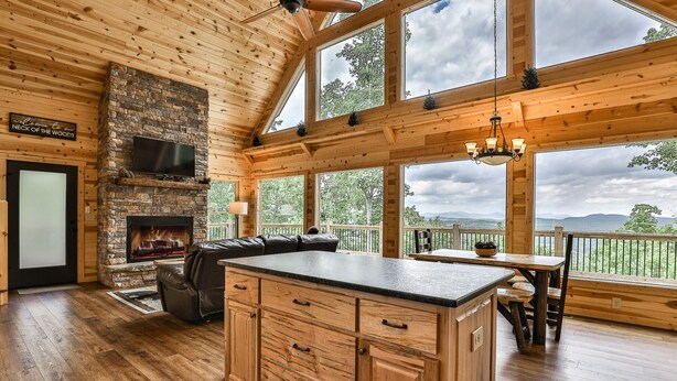 L’intérieur d’une cabane en rondins, avec une cheminée en pierre, des meubles en bois, un canapé en cuir et un coin repas offrant une vue sur l’extérieur.