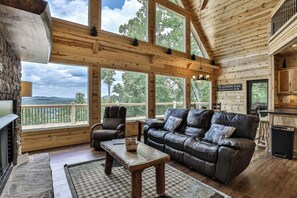 A cozy cabin interior with a fireplace, leather sofa, and large windows offering a scenic view.