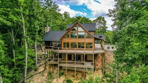 A two-story wooden cabin with a balcony, surrounded by dense forest.