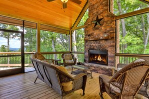 A covered deck with wicker furniture, a stone fireplace, and large windows overlooking a forest.