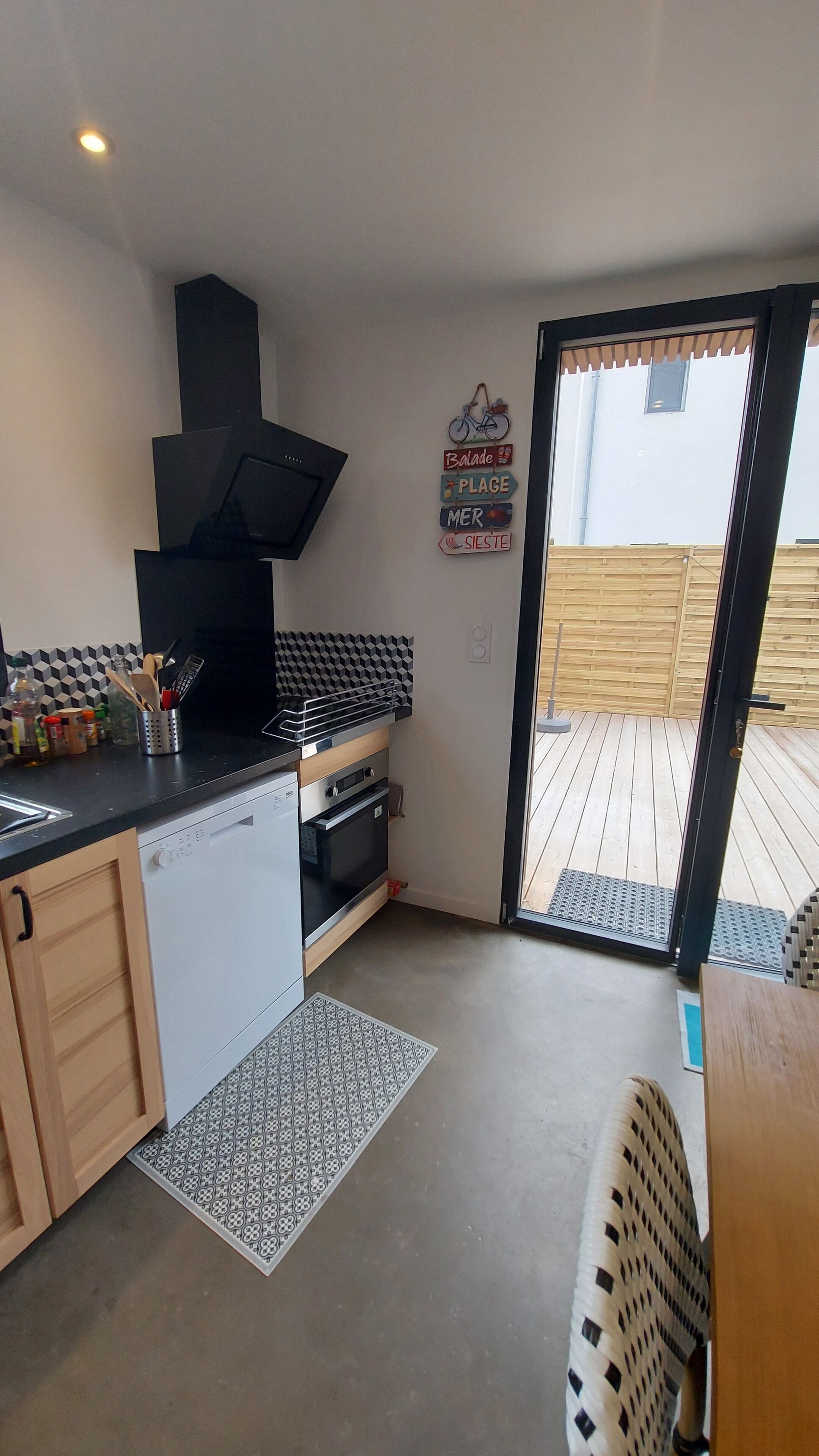 A compact kitchen with a black range hood, a built-in oven, and a dishwasher. There is a wooden cabinet with a handle, a black countertop, and a patterned rug on the floor. A bicycle sign is mounted on the wall, and a wooden deck is visible through the glass door.