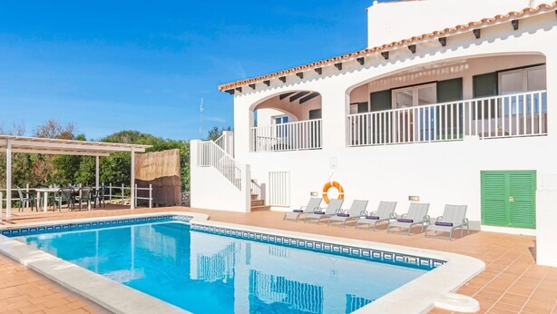 Une piscine avec des chaises longues et un espace abrité, un bâtiment blanc doté de balcons, et un ciel d’un bleu limpide.