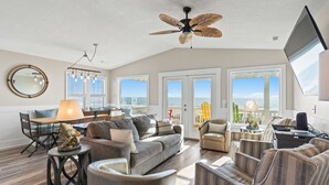 Ein Wohnzimmer mit Deckenventilator, einem runden Spiegel und Blick auf das Meer durch große Fenster.