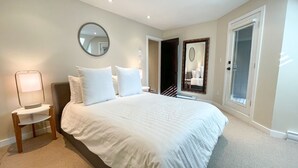 Un dormitorio con una cama grande, una mesita de noche con lámpara, un espejo redondo en la pared y una puerta que da a otra habitación.