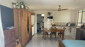 A spacious room with a large bed, a wooden wardrobe, a dining table with chairs, and a kitchen area with a refrigerator and sink. - Woody Point Coastal Cottage (Woody Point)