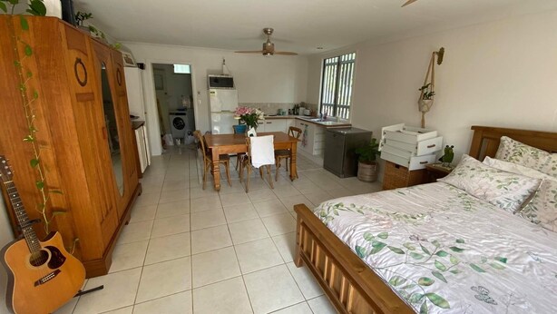A bedroom with a bed, a wooden wardrobe, a guitar, a dining table with chairs, and a kitchenette in the background. - Woody Point Coastal Cottage (Woody Point)