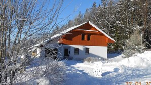 Eine zweistöckige Holzhütte mit geneigtem Dach, umgeben von schneebedeckten Bäumen und blauem Himmel.