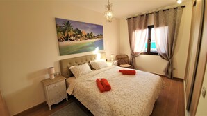 Une chambre à coucher avec un grand lit, une table de chevet, une chaise, un tableau représentant une scène de plage et une fenêtre avec des rideaux.