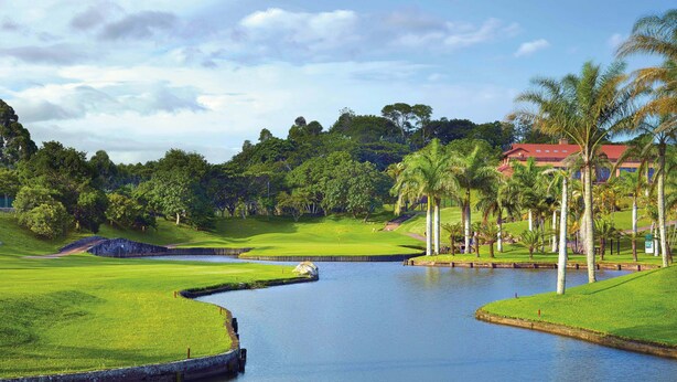 A golf course with a water feature, palm trees, and a building in the background. - San Lameer Villa Rentals 1918 (Southbroom)