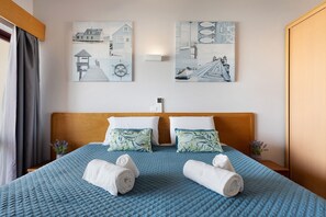 Une chambre d’hôtel avec une tête de lit en bois, deux lits avec une literie bleue, deux oreillers à motifs tropicaux et deux serviettes enroulées posées sur le lit.