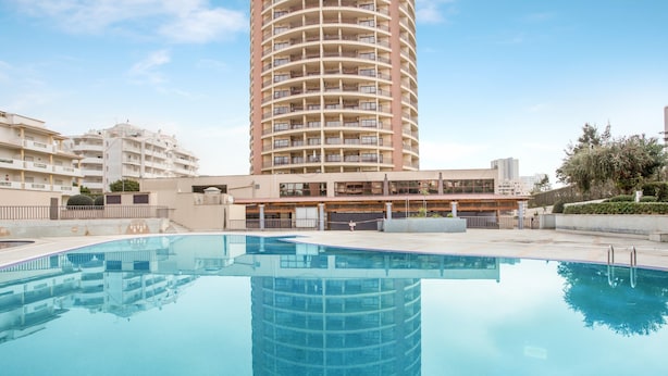 A swimming pool with a reflection of a tall building, a covered area with a roof, and adjacent residential buildings. - Apartment 'Sunny Escape' with Sea View, Wi-Fi and Air Conditioning (Portimão)