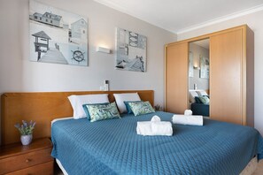Ein Schlafzimmer mit einem hölzernen Kopfteil, einem Bett mit blauer Bettwäsche, einem Nachttisch mit einer Pflanze und Wandbildern mit maritimen Motiven.