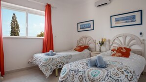 A bedroom with two beds, a window with curtains, a wall-mounted air conditioner, and two framed pictures. - Apartment "Ariana" with Pool, Wi-Fi, Balcony, Indoor Pool & Tennis Court (Portimão)