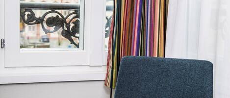 Una habitación con vistas a un edificio a través de una ventana, una silla azul, una mesa blanca con un cuenco de fruta y una lámpara negra.