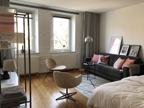 Ein modernes Hotelzimmer mit grauem Sofa, einem weißen Sessel, einem Bett, einem Schreibtisch mit Lampe und einem Fenster mit Vorhängen.