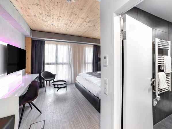A modern hotel room with a wooden ceiling, a flat-screen TV, a desk with a chair, a bed, and a bathroom with a towel rack. - Dormero HoHo Wien (Vienna)