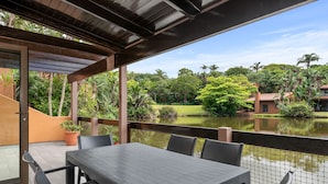 Eine Außenpatio mit Tisch und Stühlen, eine Holzpergola und Blick auf einen Teich mit umgebender Bepflanzung.