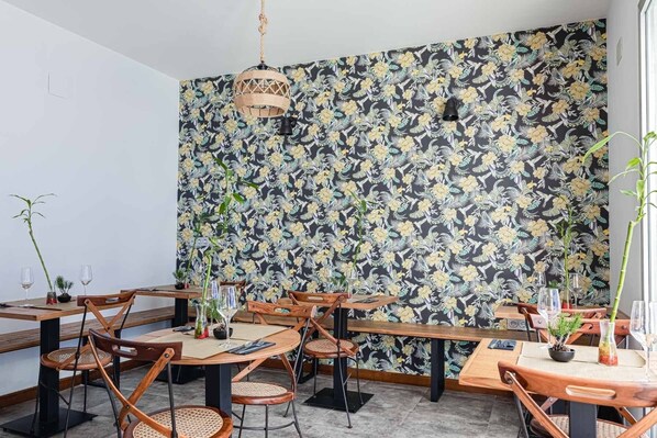 Un restaurante con mesas y sillas de madera, papel tapiz floral negro y lámparas colgantes.