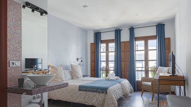 A modern bedroom with a large bed, a TV, a desk with a computer, and a bathroom with a sink and mirror. - EL COLIBRÍ RURAL HOUSE  (Sanlúcar de Guadiana)