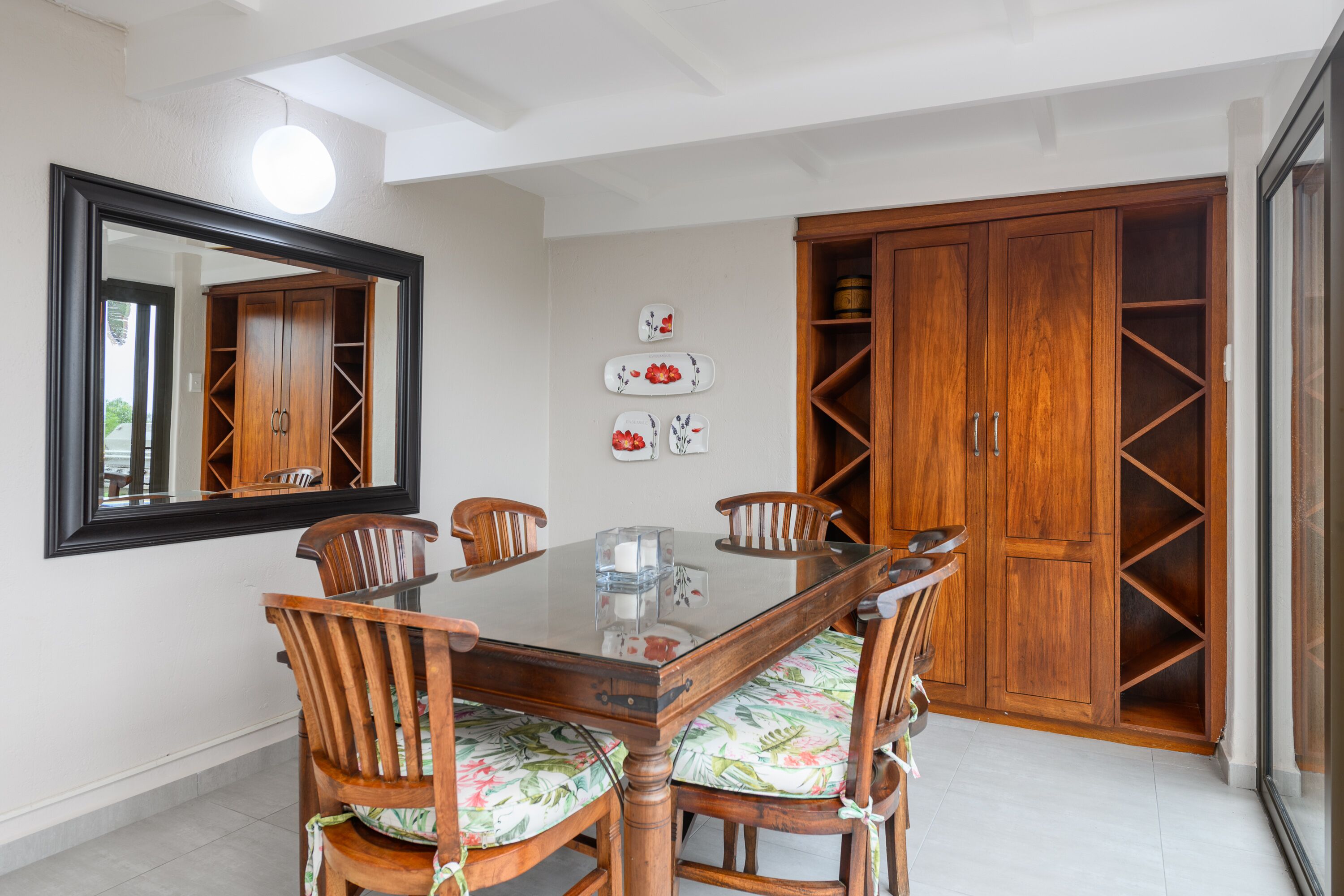 A dining area with a glass-top table, wooden chairs with floral upholstery, a large mirror, and wooden cabinets.