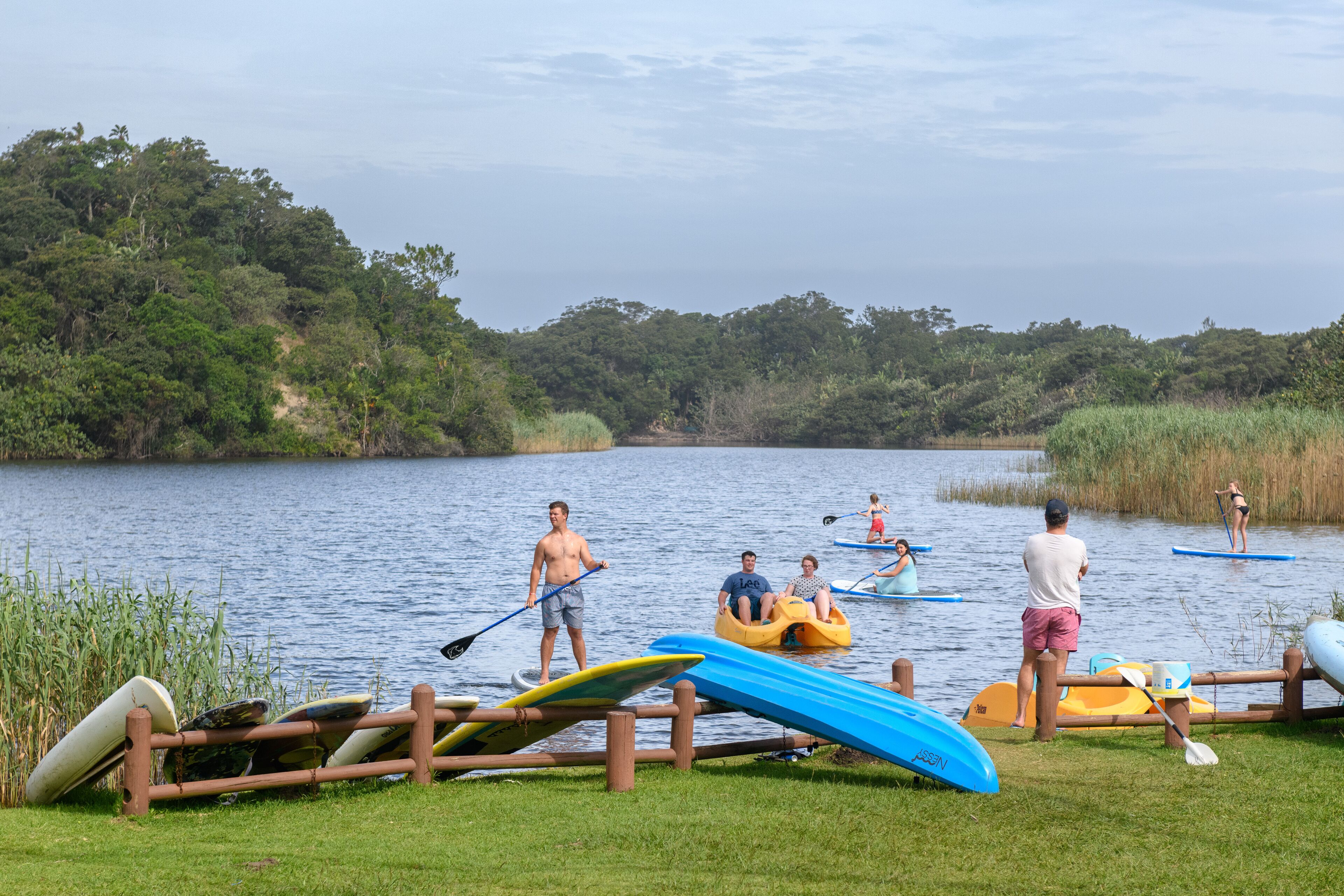 A lakeside scene with paddleboards, a wooden fence, and people engaging in water activities.