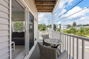 A covered balcony with a table and chairs, a sliding glass door leading to a room, and a view of a street with houses and greenery. - Beachside Abode 2A (Old Orchard Beach)