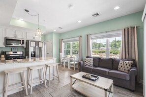 A modern kitchen with white cabinets, stainless steel appliances, and a bar area. There is a dining area with a table and chairs, and a living room with a sofa and a coffee table. - Beachside Abode 2A (Old Orchard Beach)