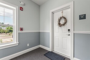 A hotel room with a white door, a fire alarm, a suite sign, a window with a view of greenery, and a doormat.