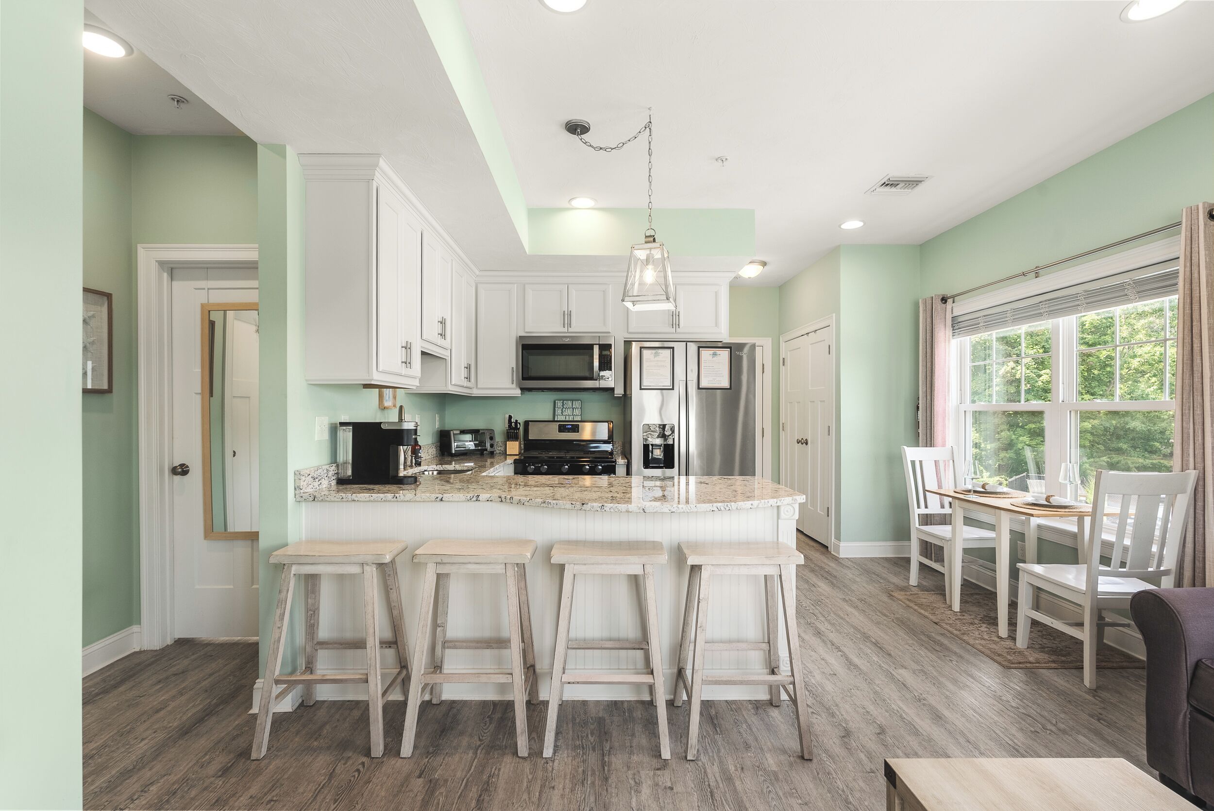 A kitchen with white cabinets, a central island with bar stools, a hanging light fixture, and a window with curtains.