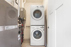 A laundry room with two stacked front-loading washing machines, a water heater, and a door. - Beachside Abode 2A (Old Orchard Beach)