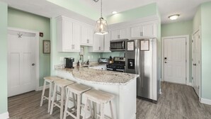 A kitchen with white cabinets, a granite countertop, a stainless steel refrigerator, and a bar stool seating area. - Beachside Abode 2A (Old Orchard Beach)