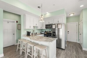 A kitchen with white cabinets, a granite countertop, a stainless steel refrigerator, and a bar stool seating area. - Beachside Abode 2A (Old Orchard Beach)