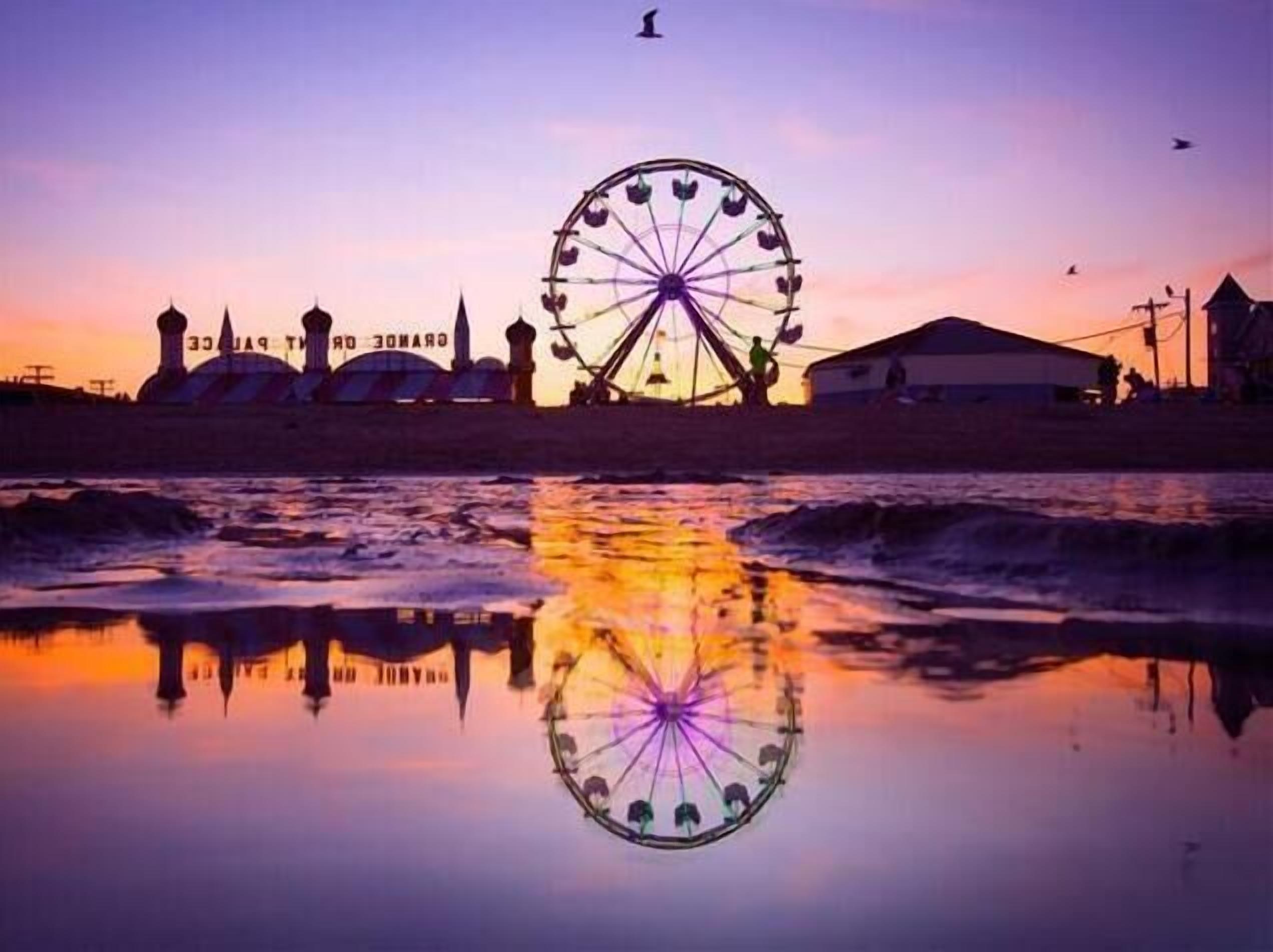 A Ferris wheel at sunset with a reflection in the water, a mosque, and a building labeled "GRAND HOTEL".